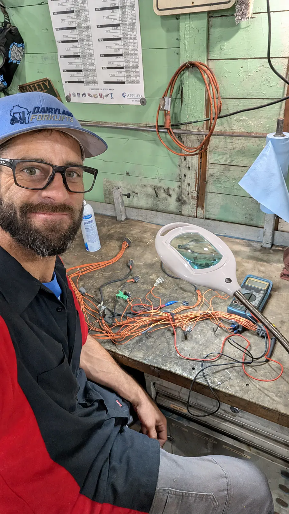 Man wearing a Dairyland Forklifts cap beside wiring and test equipment at the shop workbench